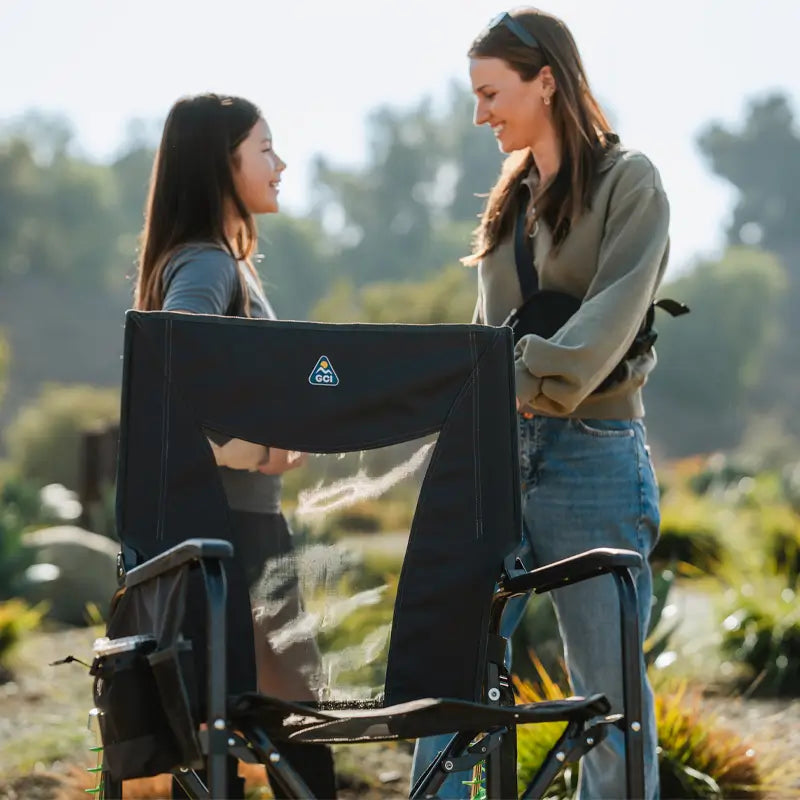 Two women chat near a black Adjustable Rocker, with trees and hills in the background.