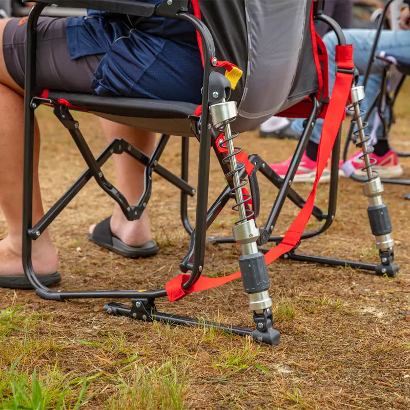 Close-up of the shock-absorbing legs and frame of a red-trimmed Adjustable Rocker in use on dry grass.