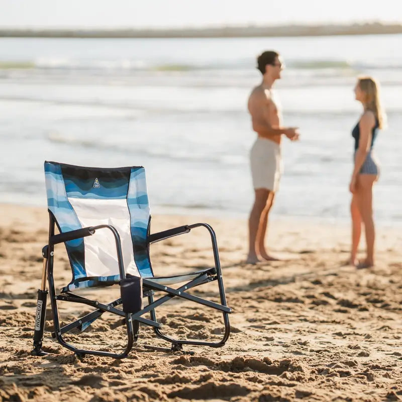Blue Beach Rocker Chair with a wave pattern sitting empty on the sand, with two people talking in the background.