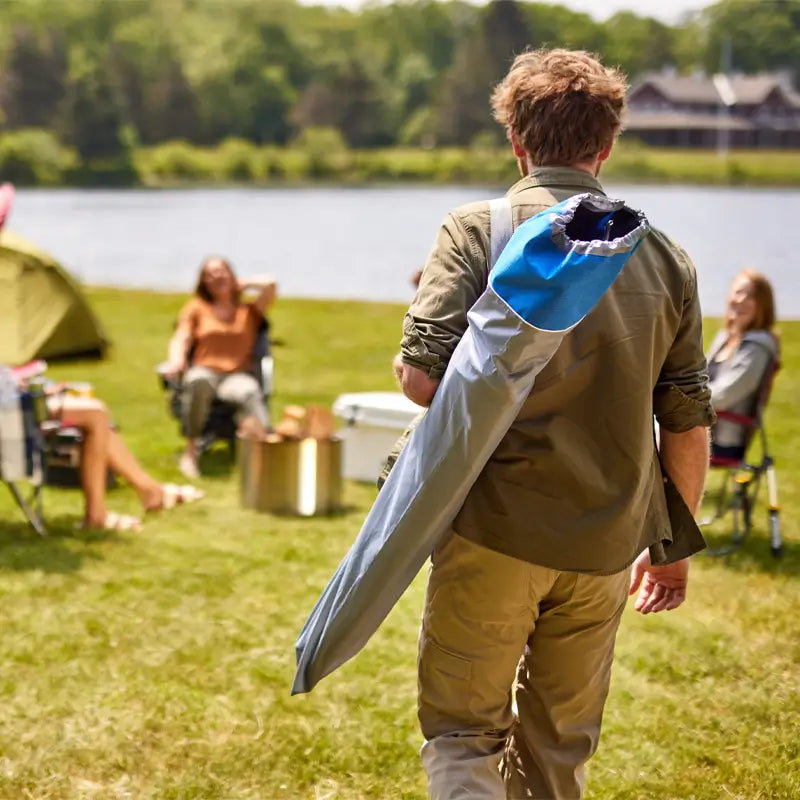 Man carrying a folded heathered royal Comfort Pro Chair in a blue and gray shoulder bag across a grassy campsite.