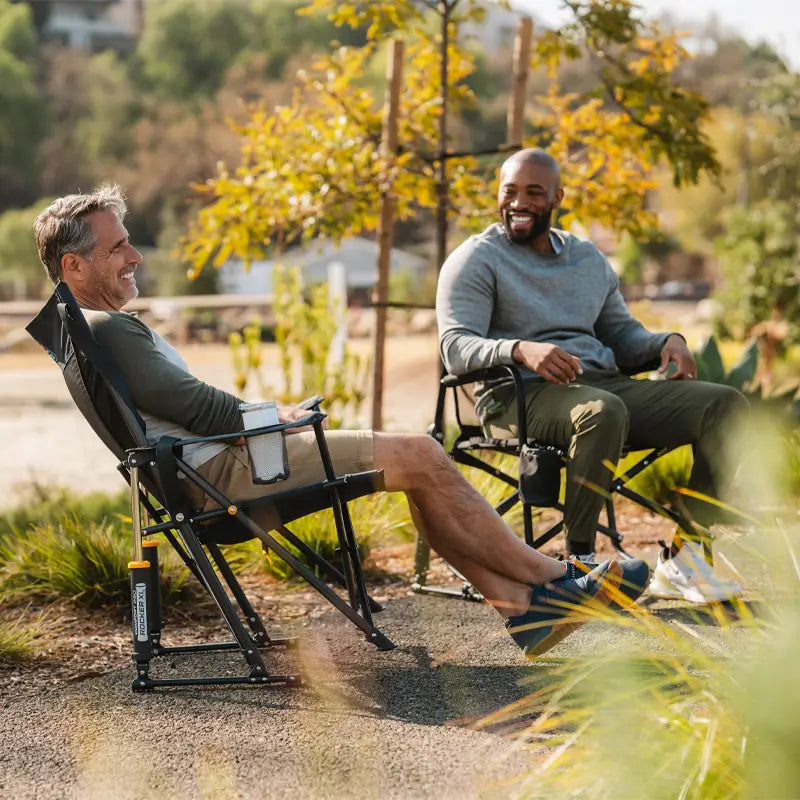 Two men laughing and relaxing in Heathered Charcoal Comfort Pro Rocker XL chairs on a scenic trail.