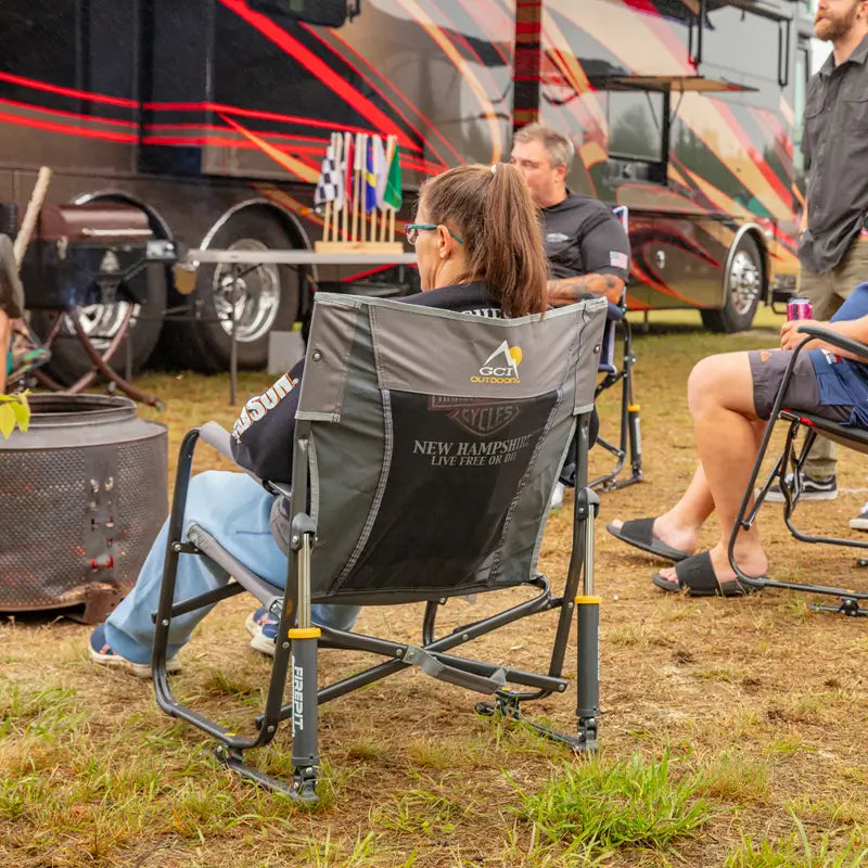 Woman relaxing in a pewter Firepit Rocker near an RV at a campsite gathering.