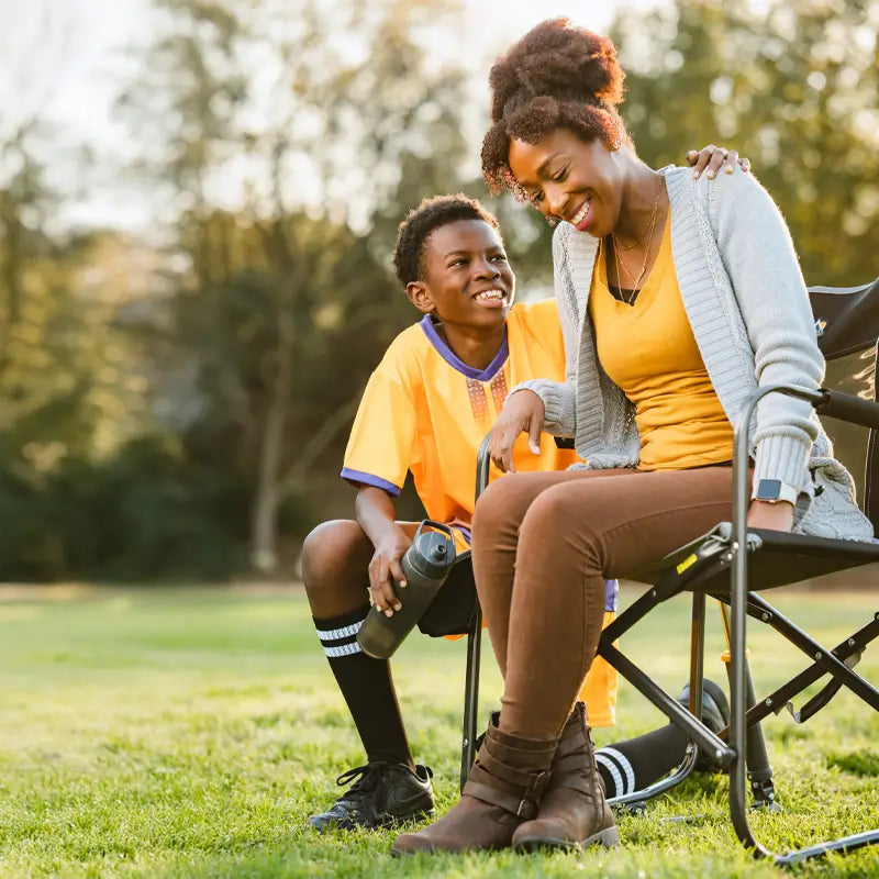 Smiling woman sits in a black Freestyle Rocker as a young soccer player in yellow jersey chats beside her after a game.