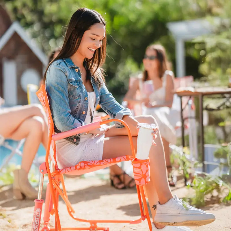 Woman in a jean jacket laughs while seated in a bold orange Freestyle Rocker with a tumbler in the holder.