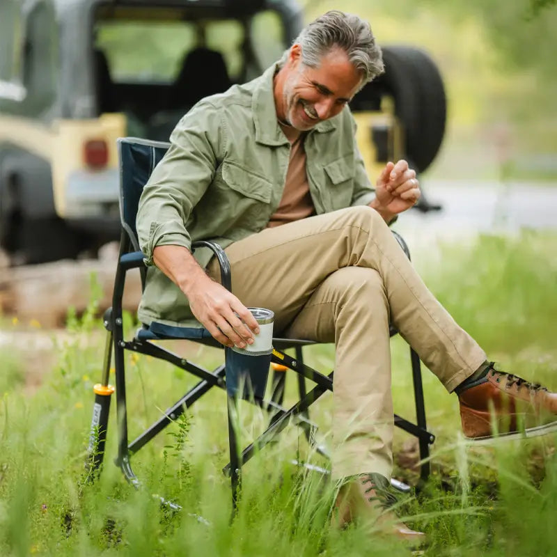A man relaxes in a Freestyle Rocker in a grassy area, holding a drink with an off-road vehicle in the background.