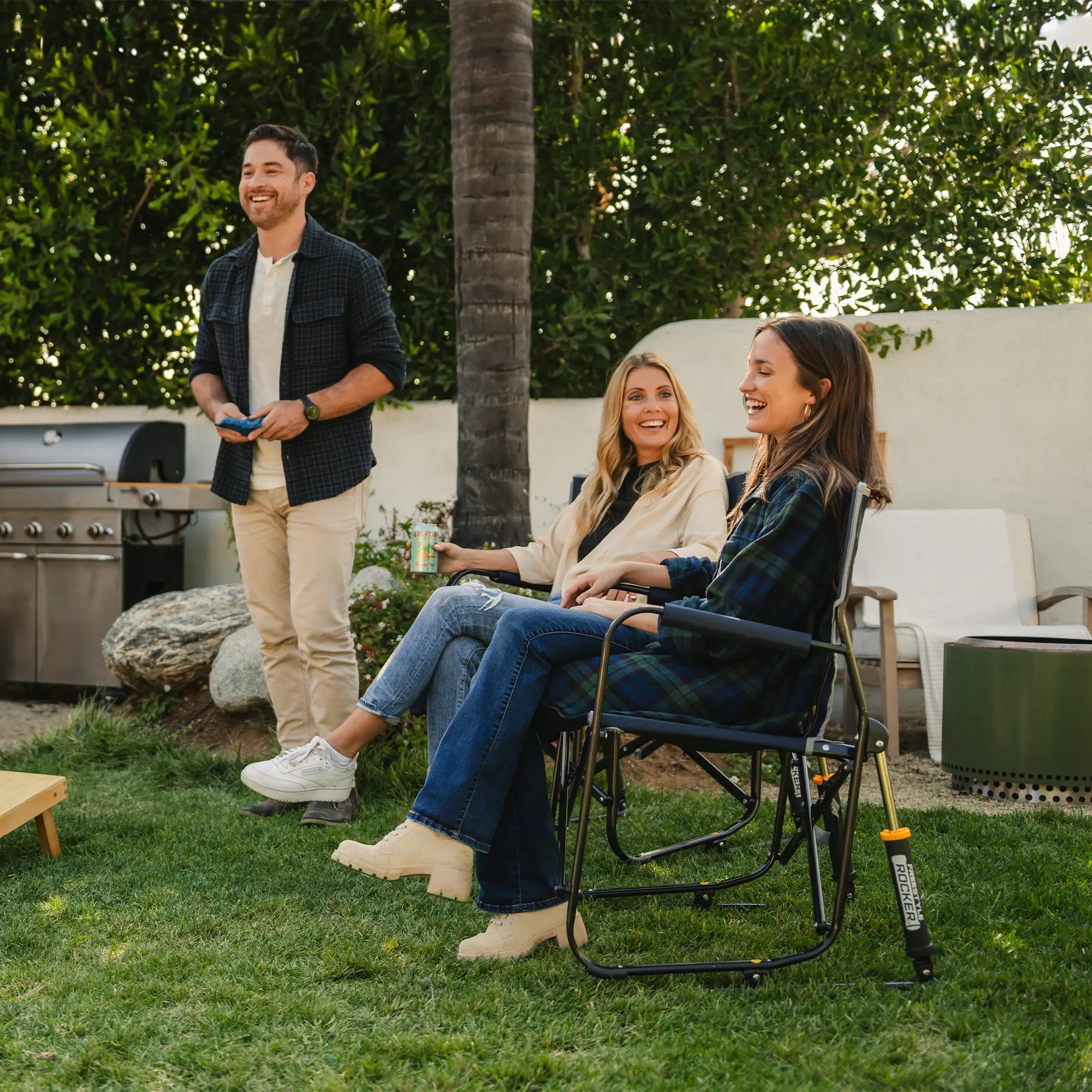 Two women watching a man play corn hole while sitting in their freestyle rocker chairs.