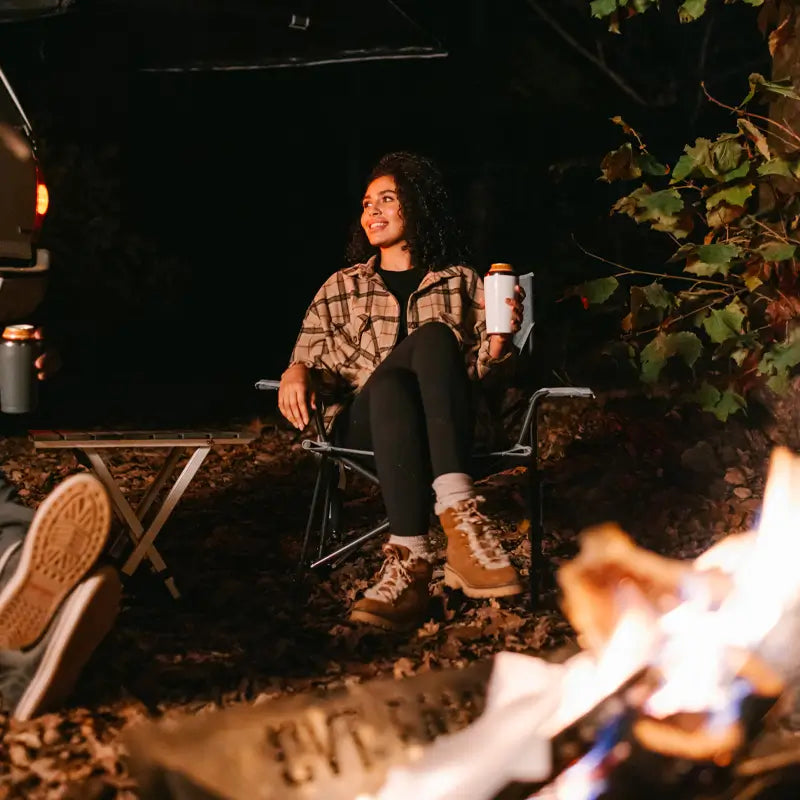 Woman enjoying a drink by a campfire, seated in the heathered pewter Kickback Rocker.