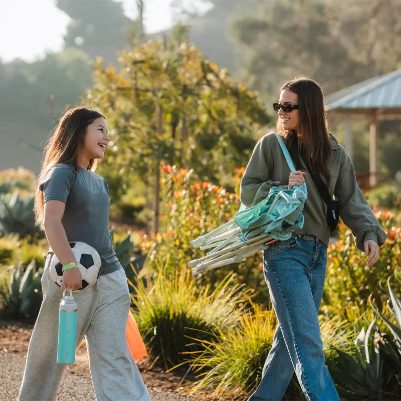 Woman carries a folded Pastel Green Leaf Kickback Rocker while walking with a girl holding a soccer ball.