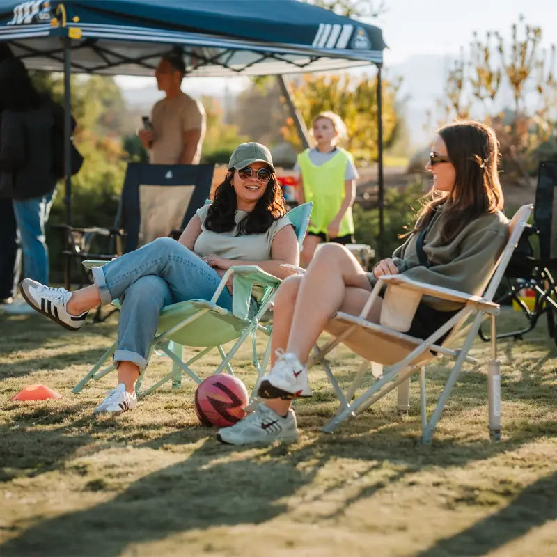 Two women chat while relaxing in pastel green leaf Kickback Rocker chairs at a youth soccer game.