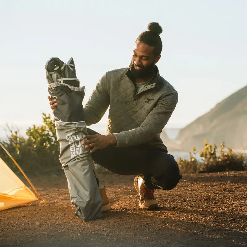 Man kneels beside the folded indigo Pod Rocker chair in its carry bag on a coastal trail. all-groups