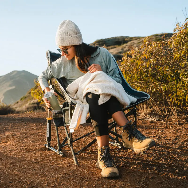 Woman relaxes in the indigo Pod Rocker chair, holding a drink with a blanket in her lap.