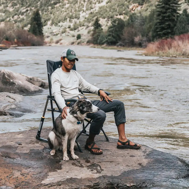 Man seated in an indigo RoadTrip Rocker on a riverside rock, resting his hand on a sitting dog while enjoying the scenic view.