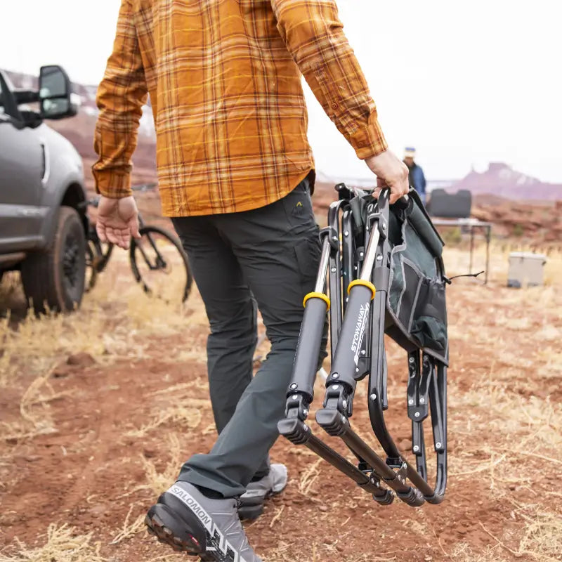 Man walking on red dirt carrying a folded hunter topo Stowaway Rocker near a vehicle.