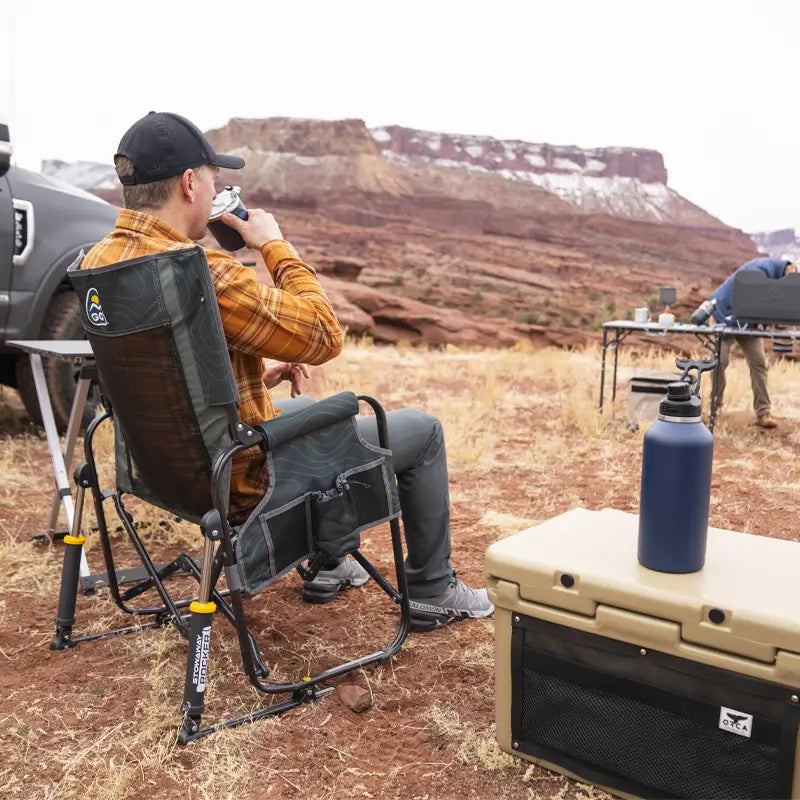 Wide shot of a man sitting in a hunter topo Stowaway Rocker at a desert campsite with a cooler nearby.