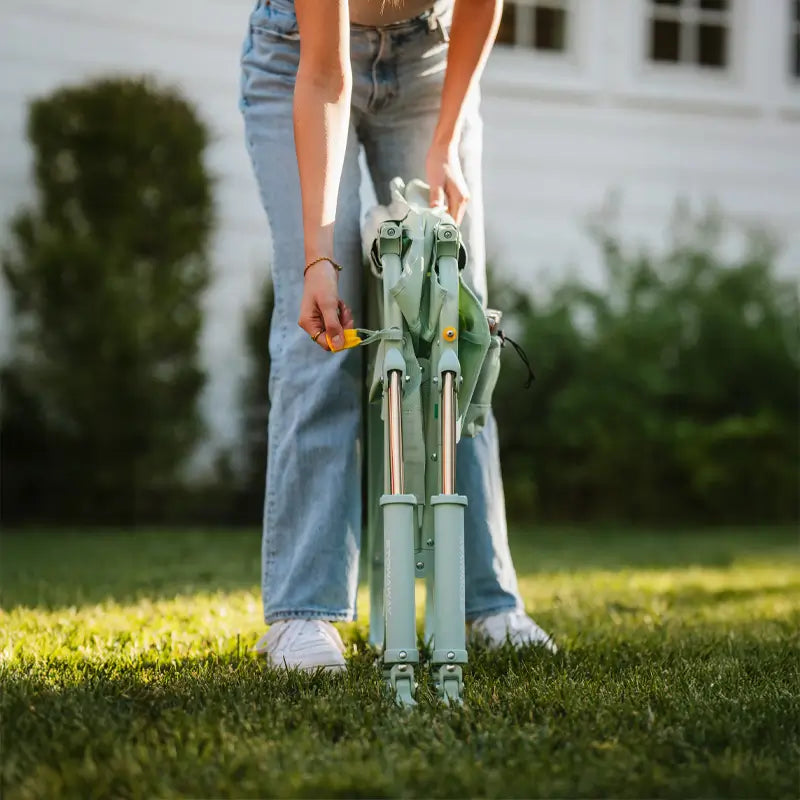 Person pulling yellow tab to unfold the pastel green Stowaway Rocker on a lawn.