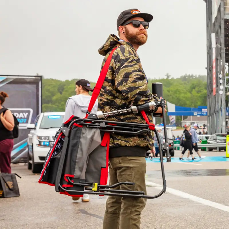 Man with a camo jacket carries a folded Adjustable Rocker using its red shoulder strap at a busy outdoor event.