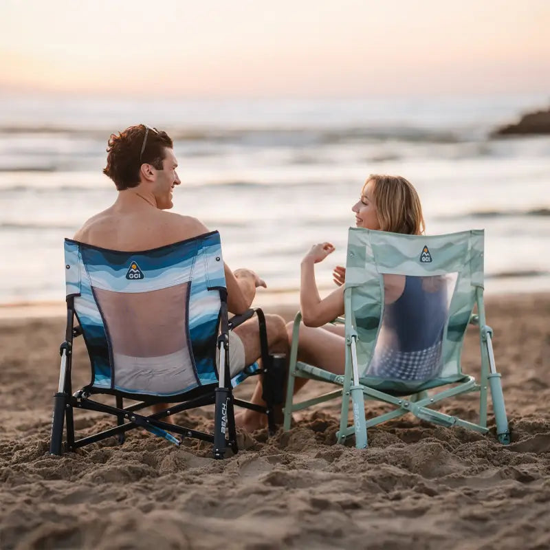 Couple sitting side by side on blue and green Beach Rocker Chairs at sunset, smiling at each other.