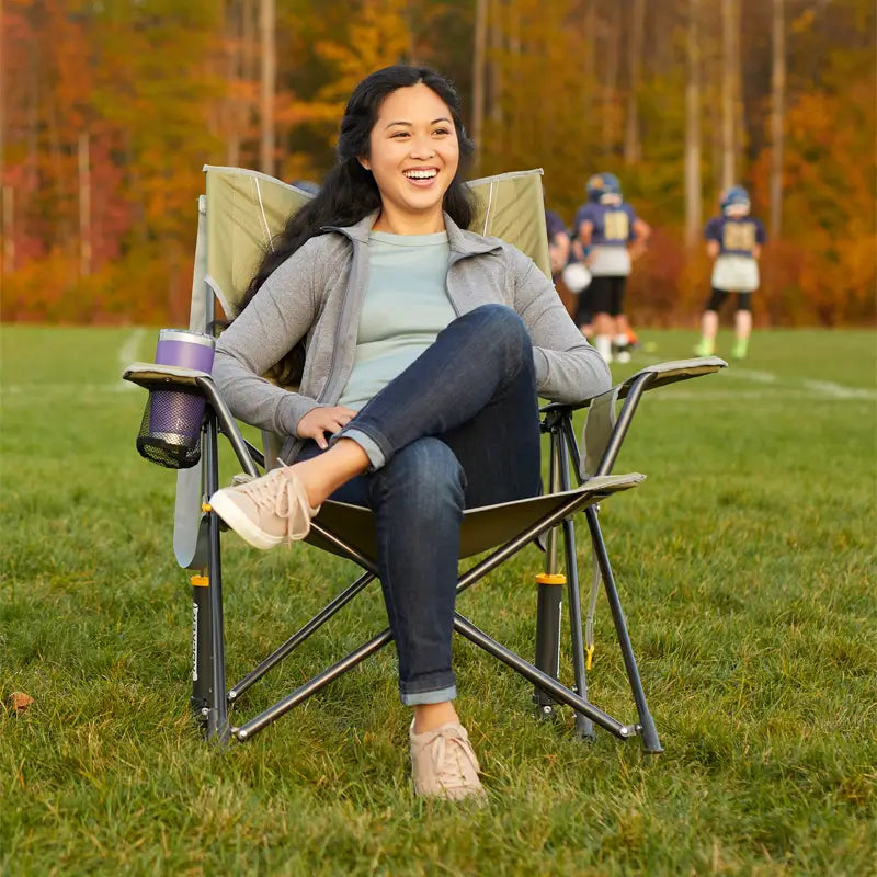 Woman smiling while seated in a GCI Comfort Pro Rocker Loden Green chair at a fall sports field with players in the background.