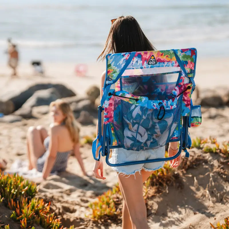 Woman carrying the folded tie dye Everywhere Chair 2 like a backpack while walking on the beach.