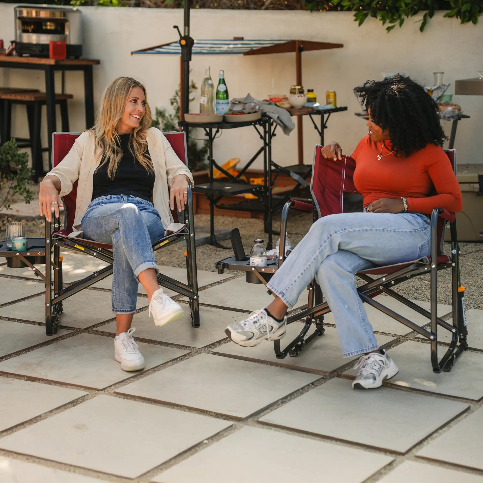 Two women conversing while sitting in their red freestyle rocker xl with side table on a patio. 