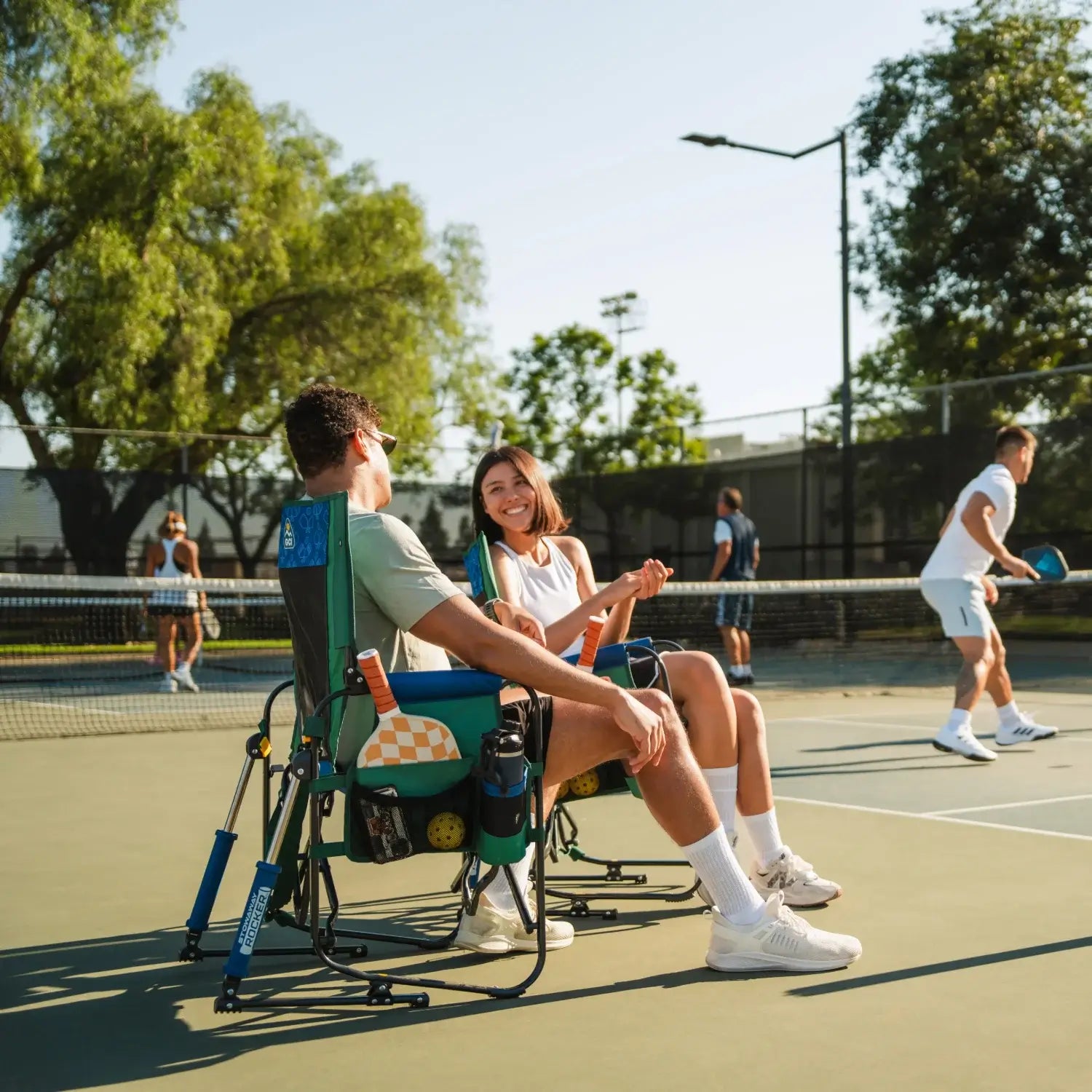 A man and woman sitting in a picklball stowaway rocker on a pickleball court. 