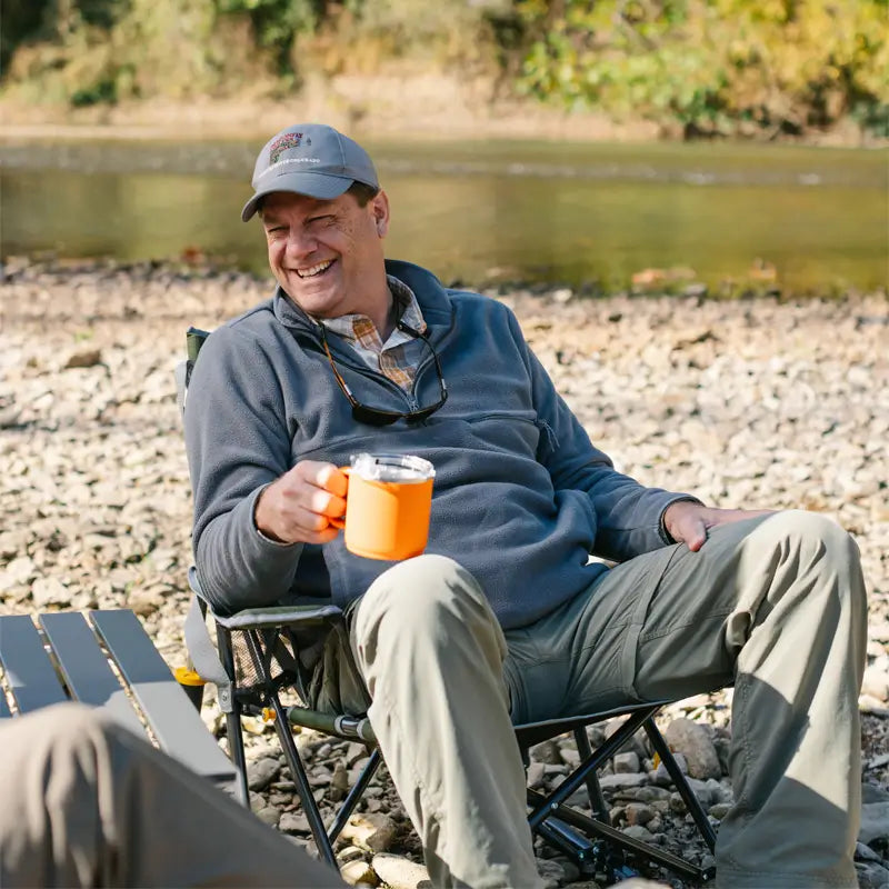 Man smiling and holding an orange mug while seated in the heathered loden Kickback Rocker by the water.