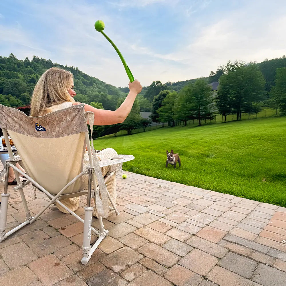 A woman throwing a tennis ball at her dog while sitting in a neutral leaf kickback rocker chair. 