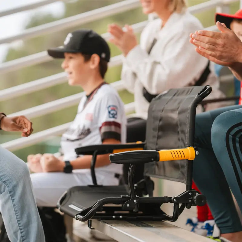 Dark charcoal Stadium Rock-Cliner positioned on bleachers beside a child in baseball uniform.