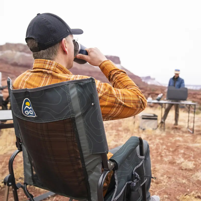 Man drinking from a tumbler while sitting in the hunter topo Stowaway Rocker at a camp setup.
