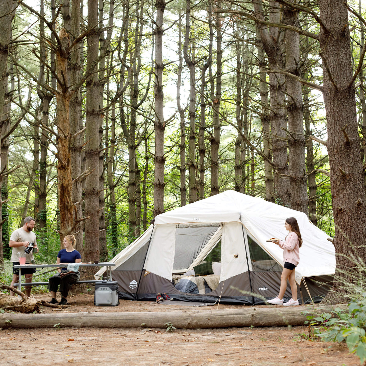 Family relaxing outdoors with CORE 10 person instant pyramid tent set up in wooded campsite, showing tall peak and space.
